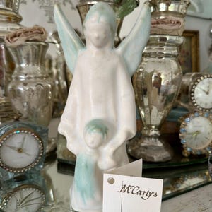 May include: A ceramic angel figurine with a matte white finish and soft blue-green accents on the wings and base. The angel holds a small child figure in front. A white paper tag attached to the base features the text McCarty's of Merigold, Mississippi.