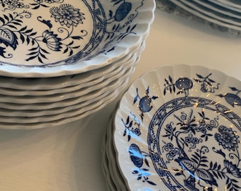 May include: Stacks of white ceramic plates with a blue floral design. The plates have a scalloped edge and a detailed pattern of flowers, leaves, and other botanical elements. The plates are stacked on a white surface.