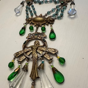 May include: A multi-strand necklace featuring a brass-toned metal figure with wings, accented by clear and green teardrop-shaped glass beads, small blue faceted beads, and two oval-shaped light green cabochons set in ornate brass-toned metal frames.