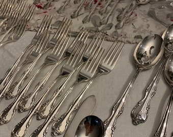 May include: A set of silver flatware, including forks, spoons, and a butter knife, arranged on a white tablecloth with a floral pattern. The flatware has an ornate design with a floral motif.