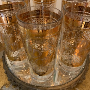 May include: Set of six gold-rimmed glass tumblers with a geometric pattern. The glasses are arranged on a round, gold-colored tray with a mirrored surface.