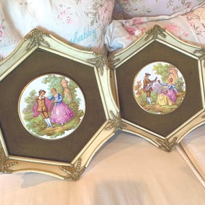 May include: Two framed vintage prints with gold accents. Each print features a round, white porcelain plate with a colorful scene of a couple in a garden setting.
