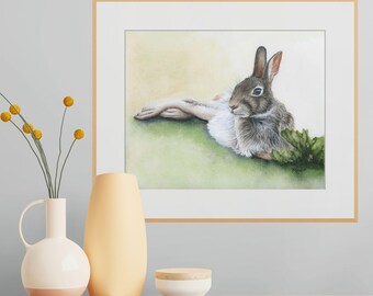 May include: A framed watercolor painting of a brown and white rabbit resting on a green surface. The rabbit is depicted in a reclining position with its ears upright. The artwork is enclosed in a light wood frame with a white mat border.