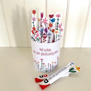 Puede incluir: Una taza de papel blanca llena de recortes con forma de flores de colores. La taza tiene texto en alemán y está decorada con un borde floral. Dos recortes florales adicionales están colocados frente a la taza. Los recortes presentan varios diseños florales.