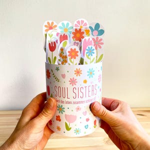 Peut inclure: Un récipient cylindrique blanc avec le texte "SOUL SISTERS" et des motifs floraux colorés. Le récipient contient de nombreuses découpes de fleurs en papier de différentes couleurs. La phrase "weil das leben zusammen schöner ist!" est également visible.