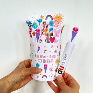 May include: A white cylindrical container decorated with colorful icons like bees, rainbows, and hearts, featuring the text "Hab einen schönen SCHULANFANG". It holds various paper cutouts and a separate stick labeled "du bist 1. Klasse".
