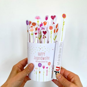 Könnte beinhalten: Ein weißer zylindrischer Behälter mit floralem Design und dem Aufdruck "HAPPY Jugendweihe", gefüllt mit dekorativen blütenförmigen Ausschnitten. Ein weißer Stab mit dem Text "bist die Heldin deiner Story" wird neben dem Behälter gehalten.