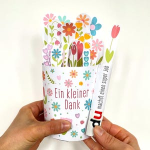 May include: A white paper flower pot with colorful floral cutouts and the text "Ein kleiner Dank". The pot holds several paper flowers in various colors. A strip of paper with the text "du machst einen super Job" is held next to the pot.