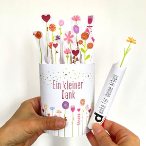 May include: A white cylindrical container decorated with colorful floral illustrations and the text "Ein kleiner Dank". Inside are several paper cutouts of flowers and hearts. One separate paper strip features a yellow flower and the text "Danke für deine Arbeit".