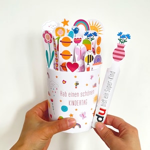 May include: A white paper cup with colorful pop-up decorations, including flowers, planets, and a rainbow. The cup has the text "Hab einen schönen KINDERTAG". A separate white card reads "du bist ein Super-Kind".