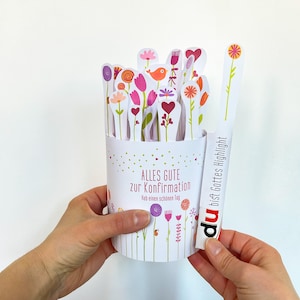 Peut inclure: Un gobelet en papier blanc avec des découpes florales et le texte "ALLES GUTE zur Konfirmation". Le gobelet contient diverses découpes de papier en forme de fleurs. Une longue bande de papier blanc avec le texte "du bist Gottes Highlight" est tenue à côté du gobelet.