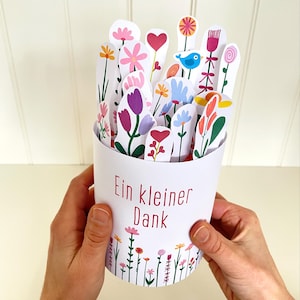 Könnte beinhalten: Ein weißer Papierbehälter gefüllt mit bunten Papierblumen, Herzen und einem blauen Vogel. Der Behälter trägt den roten Text "Ein kleiner Dank". Der Boden des Behälters hat einen floralen Rand.