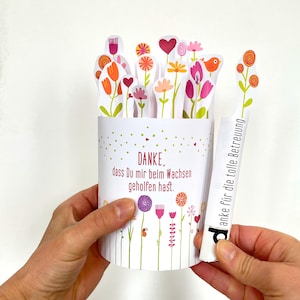 May include: A white cylindrical card with a floral design and the German text "DANKE, dass Du mir beim Wachsen geholfen hast." Colourful paper flowers and a separate card with text are held in the hands.