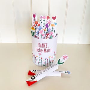 Könnte beinhalten: Ein weißer Papierbehälter gefüllt mit bunten Papierblumen. Der Behälter trägt den Text "DANKE, liebe Mami" und ein Blumendesign. Zwei papierflugzeugförmige Objekte mit floralen Akzenten und Text befinden sich vor dem Behälter.