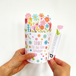 Puede incluir: Un recipiente cilíndrico blanco decorado con patrones florales coloridos y el texto en alemán "DANKE für das tolle Schuljahr". El recipiente está lleno de varios recortes de flores de papel, y una tira de papel separada dice "danke für Ihren Einsatz".