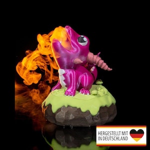 Könnte beinhalten: Eine magenta Drachenfigur, die orangefarbene Flammen ausstößt, auf einem grünen und braunen Sockel. Der Drache hat eine weiße Brust und ein rosa Horn. Das Bild enthält den Text "HERGESTELLT MIT IN DEUTSCHLAND" und eine deutsche Flagge.