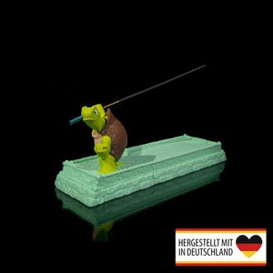 Op de afbeelding: Een grillige wierookhouder met een groene schildpadfiguur met een bruin schild, die een wierookstokje vasthoudt. De schildpad staat op een lichtgroene rechthoekige basis. De achtergrond is zwart.