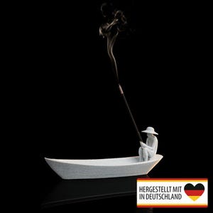 Könnte beinhalten: Ein weißer Keramik-Räucherstäbchenhalter in Form eines Bootes, mit einer Figur einer Person mit Hut, die einen Räucherstäbchen hält. Der Räucherstäbchen ist angezündet, mit Rauch, der sich vor einem schwarzen Hintergrund erhebt. Der Text "HERGESTELLT MIT IN DEUTSCHLAND" ist sichtbar.