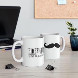 May include: Two white ceramic mugs with black text and a black mustache graphic. The mug on the left says "Firefighter Making Mustaches Great Again". The mug on the right has a black mustache graphic.