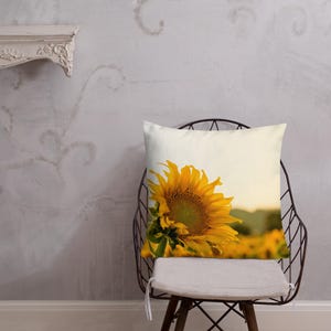 Op de afbeelding: Een kussen met een witte achtergrond en een enkele zonnebloem in het midden. De zonnebloem is naar rechts gericht en wordt gedeeltelijk bedekt door de rand van het kussen. De zonnebloem is geel met een donkerbruine kern. Het kussen ligt op een stoel met een metalen frame.