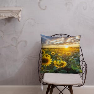 Op de afbeelding: Een kussen met een foto van een veld met zonnebloemen bij zonsondergang. De zonnebloemen staan in volle bloei en de lucht is fel oranje en geel.