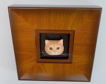 Puede incluir: Una escultura 3D enmarcada de la cara de un gato. El gato tiene pelaje naranja, ojos verdes y bigotes blancos. El marco es de madera y tiene un acabado marrón.