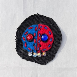 Pode incluir: Um patch de caveira feito à mão com um design dividido, metade azul e metade vermelho, bordado com fio vermelho. A caveira apresenta olhos de pedras preciosas e é colocada sobre uma base de tecido preto. O patch é embelezado com pequenas pedras preciosas iridescentes.