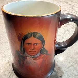 May include: A ceramic mug with a brown and black handle. The mug features a portrait of a Native American person with long dark hair and a red and white scarf. The background is a gradient of orange and red.