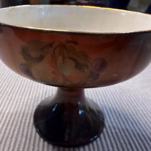 May include: A brown and gold pedestal bowl with a white interior and a painted design of grapes and leaves.