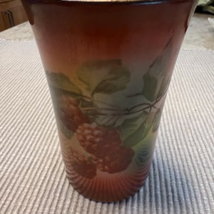 May include: A tall, tapered vase with a reddish-brown gradient. The vase features a painted design of raspberries and leaves in shades of red, green, and yellow. The rim is a light cream color.