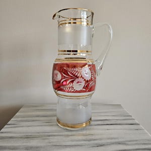 May include: A clear glass pitcher with a handle and spout, featuring frosted and red sections with gold trim. The red section is decorated with white floral designs. The pitcher sits on a marble surface.