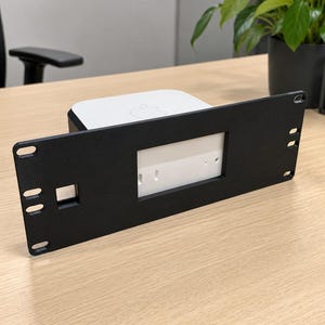 May include: A black metal rack mount bracket with a rectangular cutout and side mounting holes, designed to hold a white square electronic device with a visible Apple logo on its top surface, positioned horizontally on a flat wooden surface.