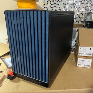 May include: A black computer case with a blue vertical-slat front panel. The case has a textured side panel and several USB ports. A spool of yellow filament sits on top. Two boxes labeled "PLA Basic" are visible.