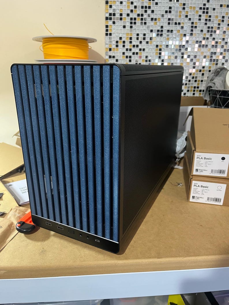 May include: A black computer case with a blue vertical-slat front panel. The case has a textured side panel and several USB ports. A spool of yellow filament sits on top. Two boxes labeled "PLA Basic" are visible.