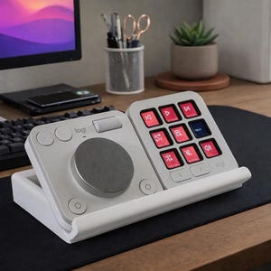 May include: A white and grey Logitech audio control device with a dial and button interface. The device has a series of red and blue illuminated buttons. The device is on a black desk mat, with a computer monitor and keyboard in the background.