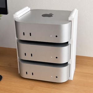 Puede incluir: Una pila de tres computadoras Mac Mini plateadas, sujetas por un soporte vertical blanco. Cada computadora tiene un diseño curvo con dos pequeños puertos negros y una luz blanca. El logotipo de Apple está en la parte superior.