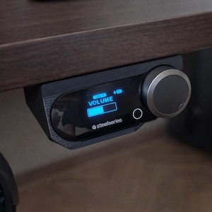 以下が含まれることがあります： 濃い茶色の机の下に取り付けられた黒いSteelSeriesオーディオコントロールデバイス。デバイスには、音量レベルを示す青色の照明付きディスプレイと大きなロータリーノブがあります。「VOLUME」という単語が画面に表示されます。