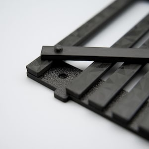 May include: Close-up of a black, geometric object with a textured surface and a series of parallel bars. A smaller rectangular piece is attached with a screw. The object is set against a white background.