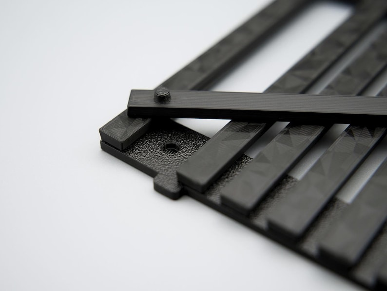 May include: Close-up of a black, geometric object with a textured surface and a series of parallel bars. A smaller rectangular piece is attached with a screw. The object is set against a white background.