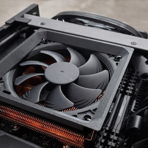 May include: Close-up of a computer's internal components, including a large, gray fan with curved blades. The fan is mounted within a dark gray frame, positioned above a copper-colored heat sink. Black cables and Corsair RAM sticks are also visible.