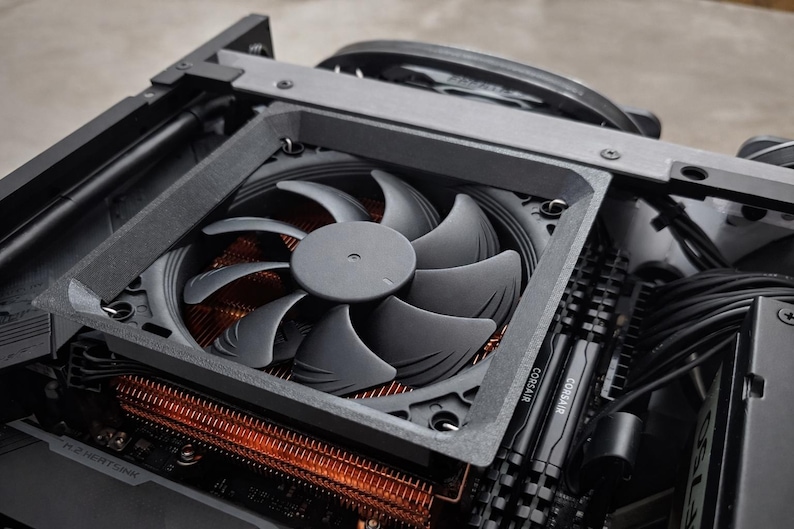 May include: Close-up of a computer's internal components, including a large, gray fan with curved blades. The fan is mounted within a dark gray frame, positioned above a copper-colored heat sink. Black cables and Corsair RAM sticks are also visible.