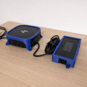 May include: A black and blue HP computer device and power supply sit on a wooden surface. The computer has a square shape with rounded corners and a blue frame. The power supply is rectangular with a blue frame.