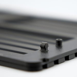 May include: Close-up of a black plastic component with a series of parallel ridges. Two small, cylindrical protrusions are visible on the surface. The object has a matte finish and appears to be part of a larger structure.