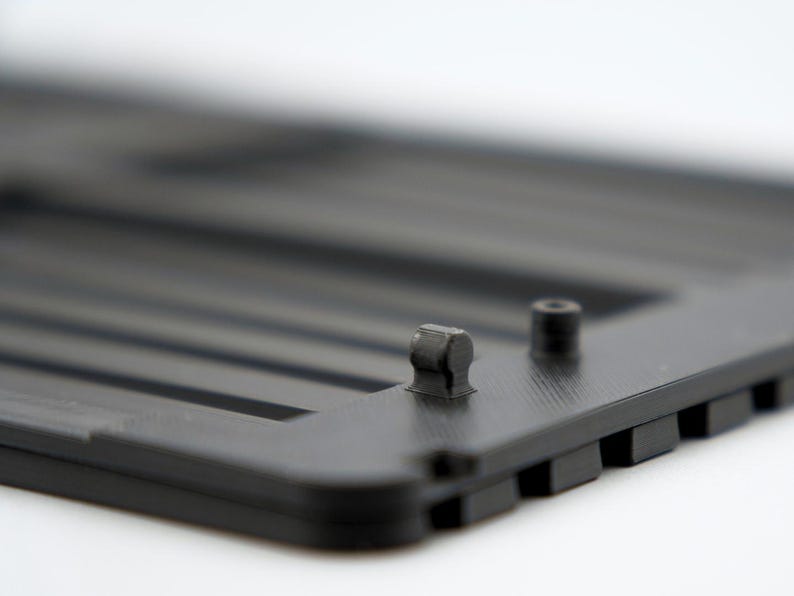 May include: Close-up of a black plastic component with a series of parallel ridges. Two small, cylindrical protrusions are visible on the surface. The object has a matte finish and appears to be part of a larger structure.