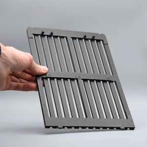 May include: A dark gray, rectangular grill grate with vertical slats. The grate is held open by a hand, showcasing its design and construction. The background is a solid, neutral gray.