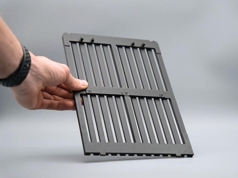 May include: A dark gray, rectangular grill grate with vertical slats. The grate is held open by a hand, showcasing its design and construction. The background is a solid, neutral gray.