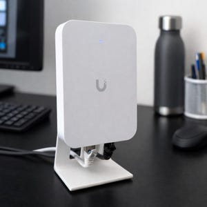 May include: A white electronic device with a rounded rectangular shape, mounted on a white stand. The device has a small blue light and a grey 'U' logo. Cables are connected to the bottom. It sits on a black desk with a keyboard, water bottle, and mouse.