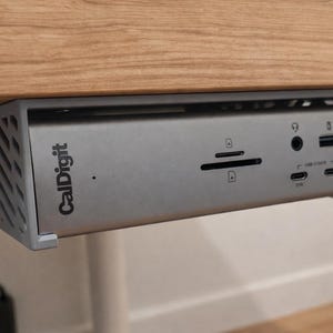 May include: A silver CalDigit docking station mounted under a wooden desk. The device has multiple ports, including USB, audio, and SD card slots. The CalDigit logo is visible on the front. The desk is supported by white legs.