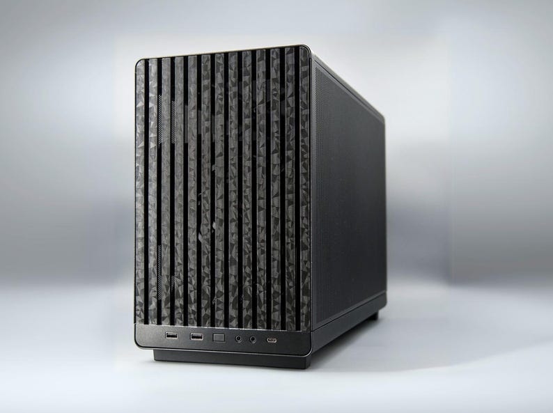 May include: A black computer case with a textured front panel featuring vertical lines and a geometric pattern. The case has a sleek, modern design with ports and buttons on the bottom.