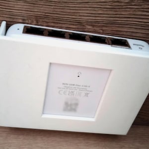 以下が含まれることがあります： 複数のポートと長方形のデザインを備えた白いネットワークスイッチ。デバイスは白いフレームに取り付けられ、木製の表面に置かれています。スイッチには小さな電源インジケーターライトとテキストのラベルが付いています。